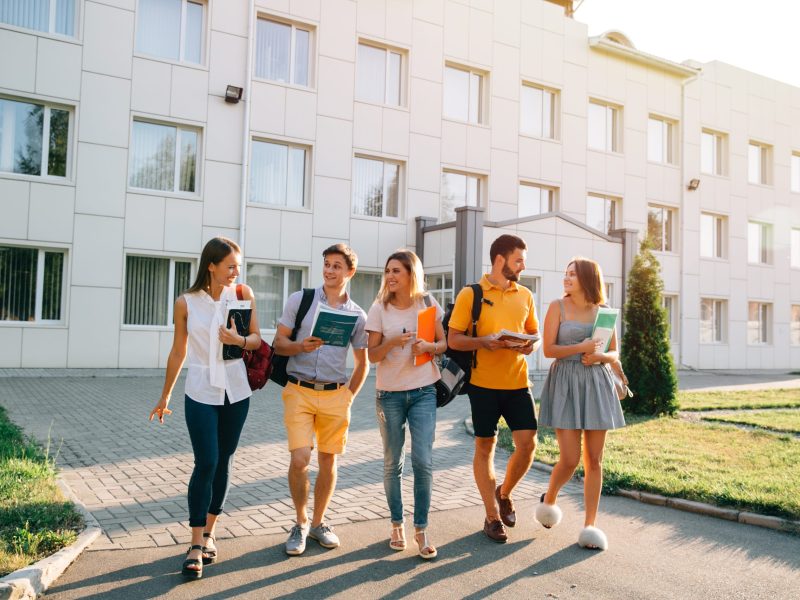 Free time of a students, bachelor`s campus life rhythm. Five friendly students are walking after they passed test outside the college building and discuss the project, smiling, enjoying, carefree